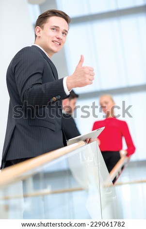 Well done. Young handsome businessman is smiling and showing OK, while standing with tablet. Two his colleagues are standing on the background and discussing important business stuff.