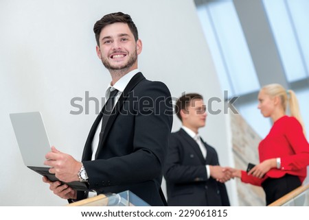 Well done. Young and motivated businessman is standing with laptop and smiling. Two his colleagues businessmen are standing on the background