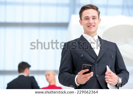 Good job. Young and motivated businessman is standing with mobile phone and tablet. Businessman is open-minded and smiling honestly