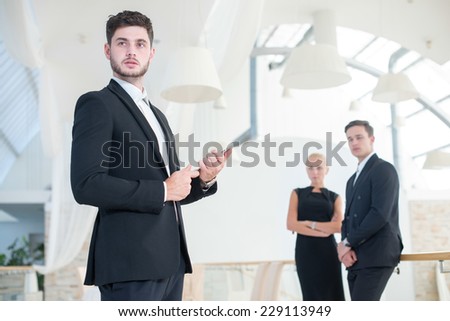 Looking in successful business future. Young motivated businessman is standing with tablet and looking forward confidently, while two his business partners are standing on the background