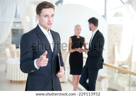 Think about this offer. Young motivated businessman is standing and looking confidently in the camera. Two his business partners are standing on the background and discussing business issues