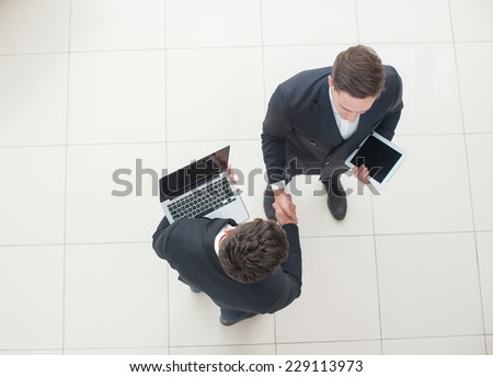 Good deal. Two young successful businessmen are standing with tablet and laptop and shaking their hands. View from the top