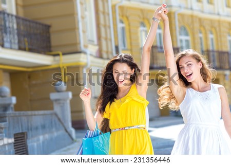 Finding the best shopping offer. Two young and pretty girls are going shopping with shopping bags. They are smiling and raising hands up