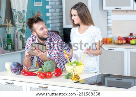 Healthy recipes in tablet on the kitchen. Beautiful pair is standing on the kitchen and cooking healthy dinner. Young  man is looking for the tasty recipes in table while his wife is making salad