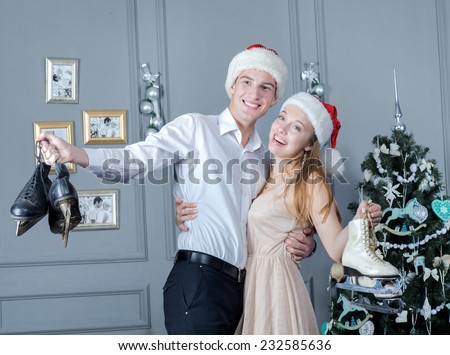 Merry Christmas. Young and beautiful pair is having great time in festive New Year living room. Both are standing in New Year Santa hats and holding figure skates. Ideal Christmas and New Year