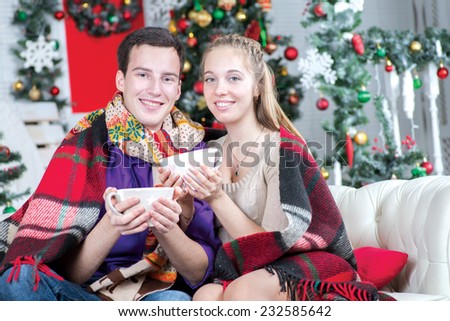 Ideal warm Christmass. Young and beautiful pair is sitting on the sofa in festive New Year living room with cups of cacao just before the New Year