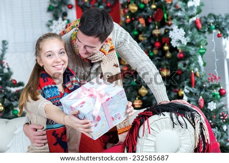 Merry Christmas and Happy New Year. Young and pretty girl is sitting with present and smiling, while her husband is standing behind her. She is showing her New Year present
