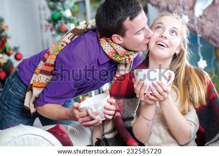 Ideal warm Christmass. Young and beautiful pair is sitting on the sofa in festive New Year living room. Young beautiful girls is holding festive cup of cacao, while her husband is standing just behind