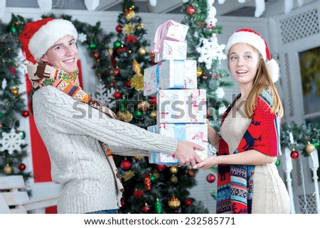 Ideal Christmas and New Year. Young and beautiful pair are standing in festive living room with presents and smiling to each other. Both are wearing New Year Santa hats