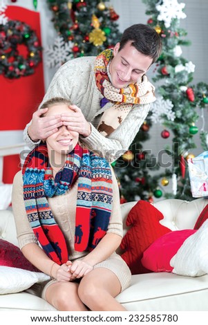 Merry Christmas.Loving couple is ready give and take Christmas presents. Young girls is sitting in festive living room with close eyes, while her husband is ready to give her New Year presents