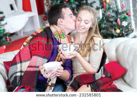 Christmas love. Young and beautiful pair is sitting on the sofa in festive New Year living room with cups of cacao and kissing each other before New Year