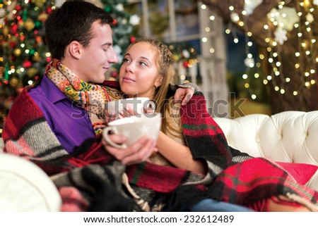 Ideal warm Christmas. Young and beautiful pair is sitting on the sofa in festive New Year living room. Pretty girl is holding beautiful festive cup of cacao
