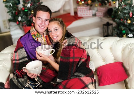 Young and beautiful pair is sitting on the sofa in festive New Year living room. Both are holding cups with New Year cacao and having festive fun