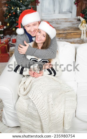 Merry Christmas and Happy New Year. Young and beautiful couple is sitting in festive New Year living room just before the Christmas. Both are drinking cacao and wearing Santa Claus hats
