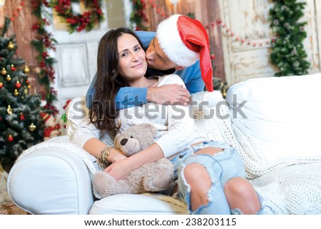 Merry Christmas and Happy New Year. Couple in love is sitting in festive Christmas decorated living room. Man is kissing his girlfriend, while she is happy just before New Year