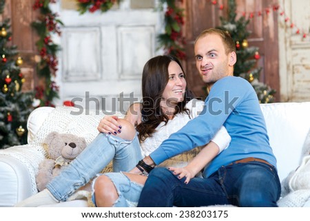 Christmas fun before New Year. Couple in love is sitting in festive Christmas decorated living room. Man is sitting with funny face, while his girlfriend is smiling and looking at him