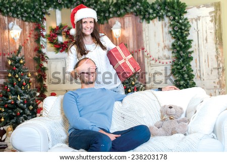 Christmas surprise. Couple in love is in festive Christmas decorated living room. Girl with smile is ready to give her husband a Christmas gift, while he is ready for New Year present.