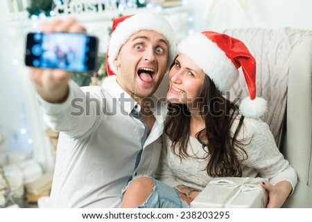 New Year funny selfie. Couple in love is in festive Christmas decorated living room is making selfie on mobile phone just before happy New Year time. Both are wearing Santa Claus hats
