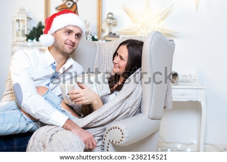 Merry Christmas and Happy New Year. Couple in love is in festive Christmas decorated living room. Both are looking at each other with happy smiles. Man is wearing Santa Claus hat