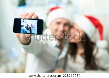Christmas selfie. Couple in love is in festive Christmas decorated living room is making selfie on mobile phone just before happy New Year time. Both are wearing Santa Claus hats