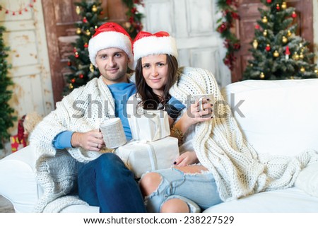 Merry Christmas and Happy New Year. Couple in love is sitting in festive Christmas decorated living room. Both are sitting with cacao in Christmas cups and looking in the camera