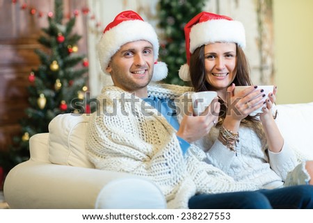 Merry Christmas and Happy New Year. Couple in love is sitting in festive Christmas decorated living room. Both are drinking New Year cacao from pretty Christmas cups and look in the camera