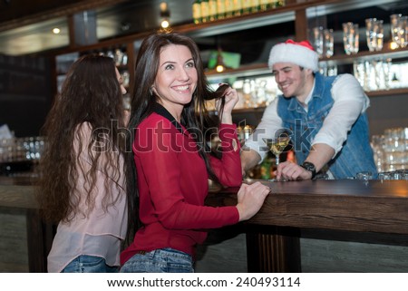 New Year in a bar. Young and beautiful girl is standing in a bar or pub and smiling. Bartender in Christmas hat is standing behind bar counter. Christmas is good with beer.
