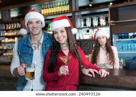 Love beer and Christmas. Loving couple is standing with beer in a bar or pub, wearing New Year Santa Claus hats. Girls bartender also is wearing Santa Christmas hat and stays on a background