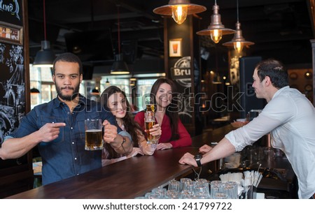 Perfect beer party. Portrait of young and handsome guy. He is standing at the bar counter and showing his beer. His female friends are standing with beer on the background and talking with each other