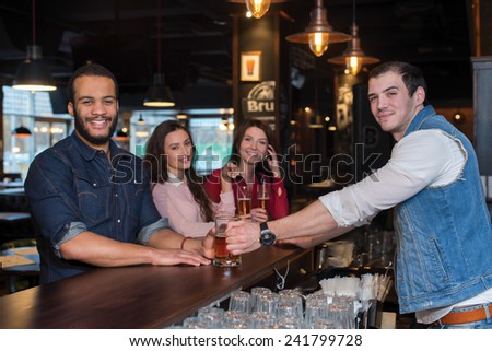 Great evening with beer. Portrait of young and handsome afro american guy. He is standing at the bar counter and talking with barman, while drinking beer. His friends are standing with beer behind