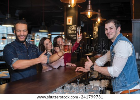 Having some fun with beer. Portrait of young and handsome afro american guy. He is standing at the bar counter and showing his beer. His female friends are standing with beer on the background
