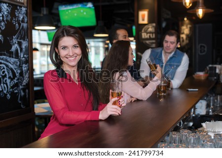 Perfect beer party. Portrait of young beautiful girl is standing in a pub with glass of beer and smiling. Her friends are standing next to her and talking with barman