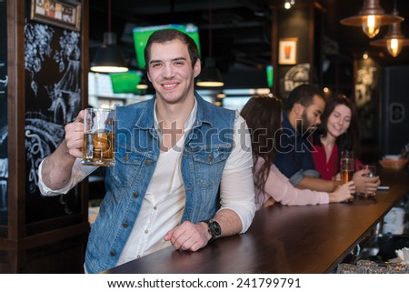 Spending time with beer. Portrait of young handsome man who is standing in a pub with glass of beer and smiling. His friends are standing on the background