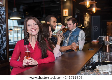 Perfect beer party. Portrait of young beautiful girl is standing in a pub with glass of beer and smiling. Her friends are standing next to her and drinking beer