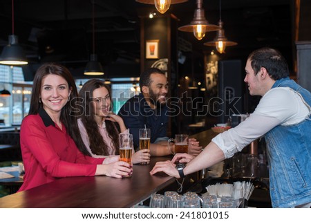 Perfect beer party. Portrait of young beautiful girl is standing in a pub with glass of beer and smiling. Her friends are standing next to her and talking with barman