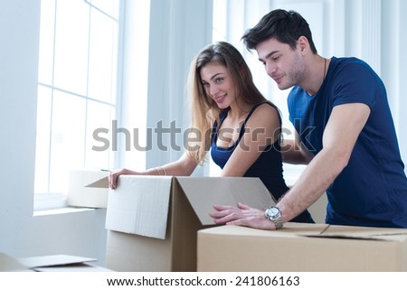 Moving new flat with fun and excitement. Young and beautiful couple is moving to new apartment surrounded with plenty of cardboard boxes. Both are opening boxes with smiles