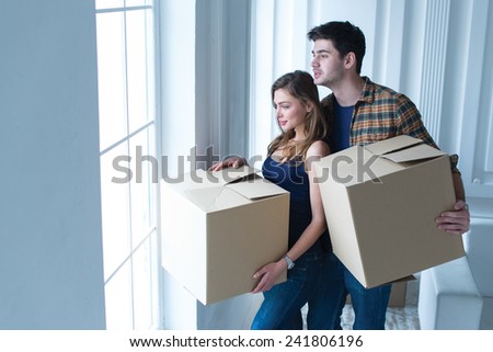 Entering new house. Young and beautiful couple is moving to new apartment surrounded with plenty of cardboard boxes. Both are looking in the window and holding boxes