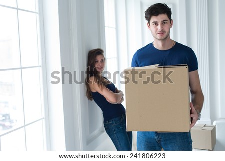 Moving new flat with fun and excitement. Portrait of young and handsome man moving to new apartment surrounded with plenty of cardboard boxes. His beautiful wife is on the background