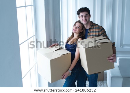 Entering new house. Young and beautiful couple is moving to new apartment surrounded with plenty of cardboard boxes. Both are standing near the window and holding boxes