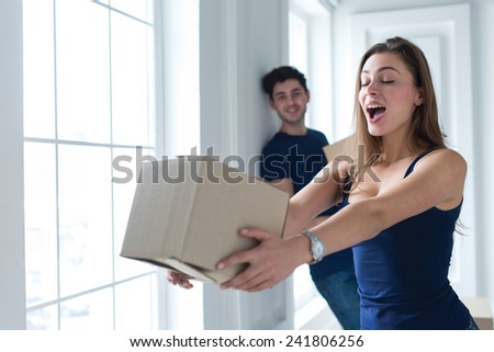 Moving new flat with fun and excitement. Portrait of young and beautiful girl moving to new apartment surrounded with plenty of cardboard boxes. Her handsome husband is on the background
