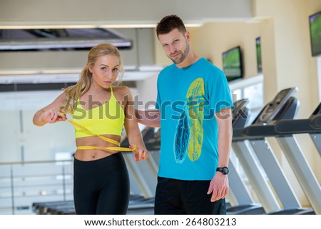 Great training result. Young and pretty woman is measuring her training result. Handsome athlete man is standing next to her and confidently looks in camera