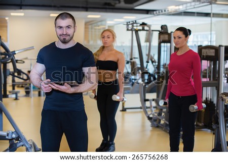 Intensive training together. Portrait of young and handsome strong athlete man in a gym. Two athlete girls are training on the background. Perfect shape and proper training.