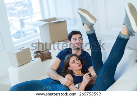 Moving new flat with fun and excitement. Young and beautiful couple is moving to new apartment surrounded with plenty of cardboard boxes. Both are sitting on the sofa and dreaming about new cozy home