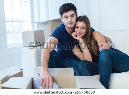 Moving new flat with fun and excitement. Young and beautiful couple is moving to new apartment surrounded with plenty of cardboard boxes. Both are sitting on the sofa and unpacking boxes