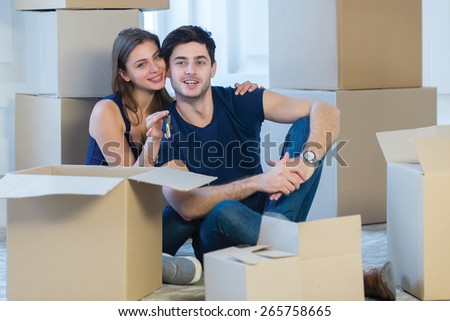 Moving new flat with fun and excitement. Young and beautiful couple is moving to new apartment surrounded with plenty of boxes. Both are sitting on the floor and holding keys for their new cozy house