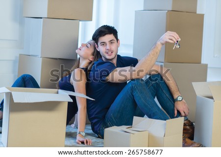 Moving new flat with fun and excitement. Young and beautiful couple is moving to new apartment surrounded with plenty of boxes. Both are sitting on the floor and holding keys for their new cozy house