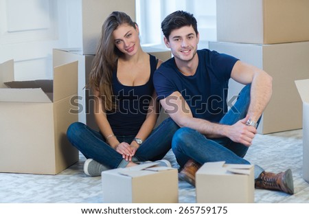 Moving new flat with fun and excitement. Young and beautiful couple is moving to new apartment surrounded with plenty of cardboard boxes. Both are sitting on floor and dreaming about new cozy house