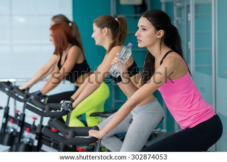 Perfect shape and ideal workout. Young and pretty woman is having training on exercise bike and drinks water. Her friends are also on exercise bikes are next to her. Active healthy workout in a gym.