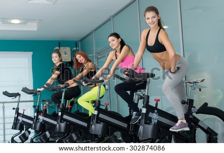 Perfect shape and ideal workout. Young and pretty woman is having training on exercise bike. Her friends are also on exercise bikes are next to her. Active workout in a gym. Healthy sportsmen concept
