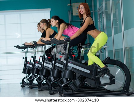 Perfect shape and ideal workout. Young and pretty woman is having training on exercise bike. Her friends are also on exercise bikes are next to her. Active workout in a gym. Healthy sportsmen concept